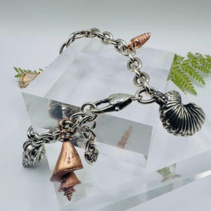 May include: A silver chain link charm bracelet featuring multiple seashell-shaped charms in silver and copper tones. The bracelet includes a heart-shaped clasp and a small dangling charm with the letter B engraved on it, showcasing detailed textures.