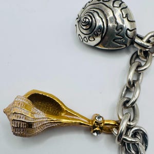 May include: A silver-toned chain bracelet featuring two distinct seashell charms. One charm is a rounded, textured silver shell, while the other is a gold-toned conch shell with white accents and two small, clear embedded crystals at the base.