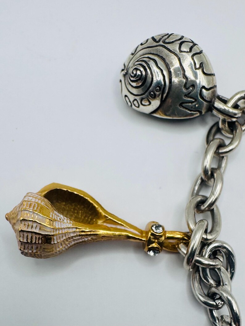 May include: A silver-toned chain bracelet featuring two distinct seashell charms. One charm is a rounded, textured silver shell, while the other is a gold-toned conch shell with white accents and two small, clear embedded crystals at the base.