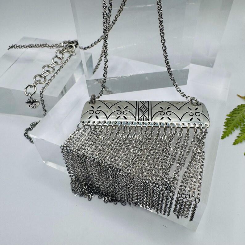 May include: A silver-toned necklace featuring a rectangular metal pendant with engraved geometric and floral patterns. A dense fringe of multiple thin silver chains hangs from the bottom edge of the pendant, suspended by a delicate silver link chain.