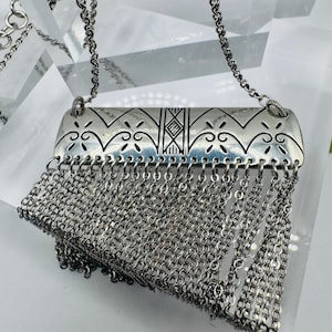May include: A silver-toned necklace featuring a rectangular metal pendant with engraved geometric and scroll patterns. The center of the pendant displays the letters HH. A dense fringe of multiple silver-toned chain links hangs from the bottom.