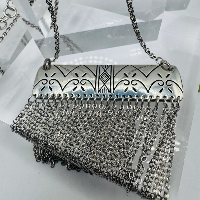 May include: A silver-toned necklace featuring a rectangular metal pendant with engraved geometric and scroll patterns. The center of the pendant displays the letters HH. A dense fringe of multiple silver-toned chain links hangs from the bottom.