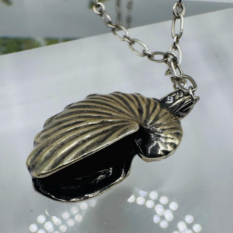 May include: A silver-toned metal pendant shaped like a nautilus shell, featuring deep ridges and a dark, oxidized finish. The pendant is attached to a silver-toned chain link necklace and includes a small, clear, sparkling stone accent near the top.