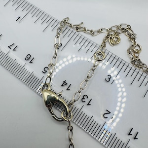 May include: A silver-toned metal chain necklace featuring a heart-shaped lobster claw clasp. The necklace is positioned next to a clear plastic ruler showing measurements in inches, with the clasp measuring approximately 0.5 inches in length.
