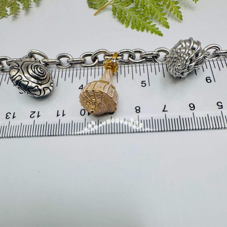 May include: A silver-toned chain bracelet featuring three distinct charms: a textured silver snail shell, a gold-toned textured conical shell, and a detailed silver nautilus shell. A ruler is positioned behind the charms, showing a scale in centimetres.