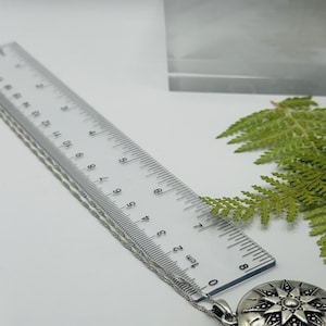 May include: A silver-toned circular pendant necklace featuring a starburst design with dark accents. A clear plastic ruler is positioned alongside the chain, showing the pendant measures approximately 1.25 inches in diameter.