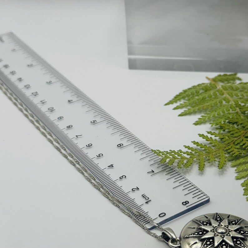 May include: A silver-toned circular pendant necklace featuring a starburst design with dark accents. A clear plastic ruler is positioned alongside the chain, showing the pendant measures approximately 1.25 inches in diameter.