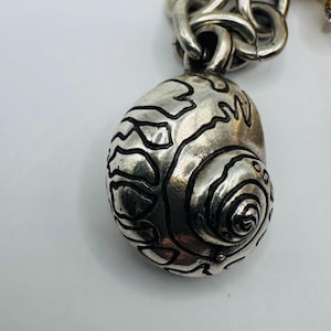 May include: A silver-toned metal charm shaped like a spiral seashell with dark, recessed etched lines detailing the shell's surface. The charm is attached to a thick, interlocking silver-toned metal chain link bracelet.