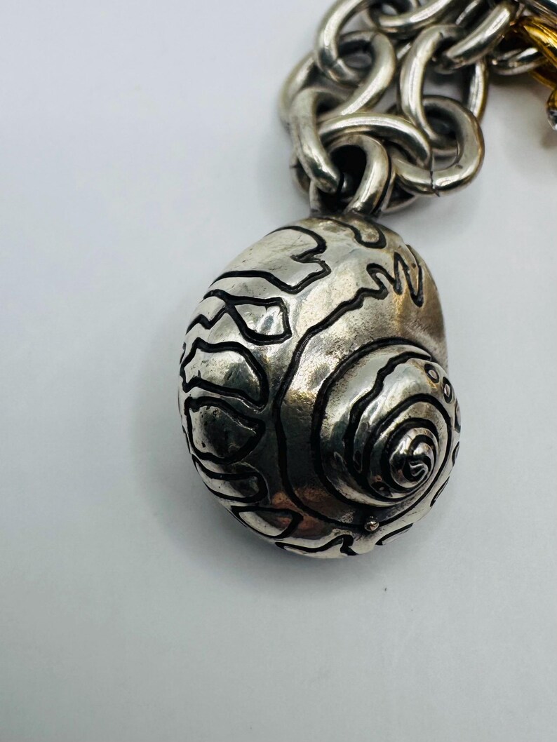 May include: A silver-toned metal charm shaped like a spiral seashell with dark, recessed etched lines detailing the shell's surface. The charm is attached to a thick, interlocking silver-toned metal chain link bracelet.
