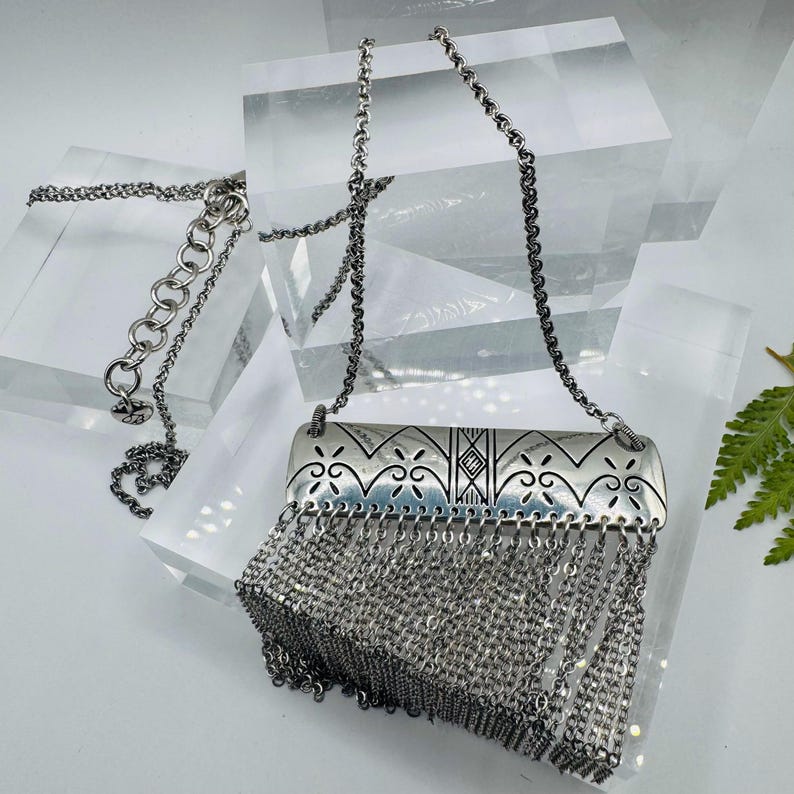 May include: A silver-toned necklace featuring a rectangular metal pendant with engraved geometric and scroll patterns. The pendant is adorned with a dense fringe of multiple thin silver chains hanging from the bottom edge, attached to a link chain.