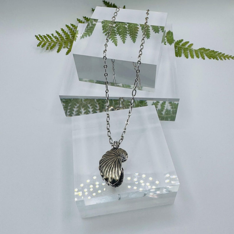 May include: A silver-toned necklace featuring a pendant shaped like a textured seashell. The pendant hangs from a delicate silver-toned metal chain with oval links. The design captures the natural ridges and curved form of a shell in metallic silver.