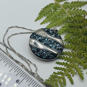 May include: A circular silver-toned pendant necklace featuring three diagonal bands of small, sparkling blue crystals. The pendant is attached to a thin silver-toned chain, and a ruler is visible showing a diameter of approximately 1.5 inches.