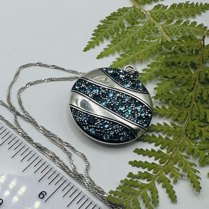 May include: A circular silver-toned pendant necklace featuring three diagonal bands of small, sparkling blue crystals. The pendant is attached to a thin silver-toned chain, and a ruler is visible showing a diameter of approximately 1.5 inches.
