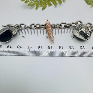 May include: A silver-toned metal chain bracelet featuring three distinct charms: a dark nautilus shell, a central rose gold-toned spiral shell, and a patterned round shell. A ruler is positioned beneath the charms, showing a length of 20 cm.