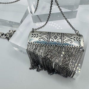 May include: A silver-toned necklace featuring a rectangular metal pendant with engraved symmetrical scrollwork and geometric patterns. A dense fringe of multiple thin silver-toned metal chains hangs from the bottom edge of the pendant.