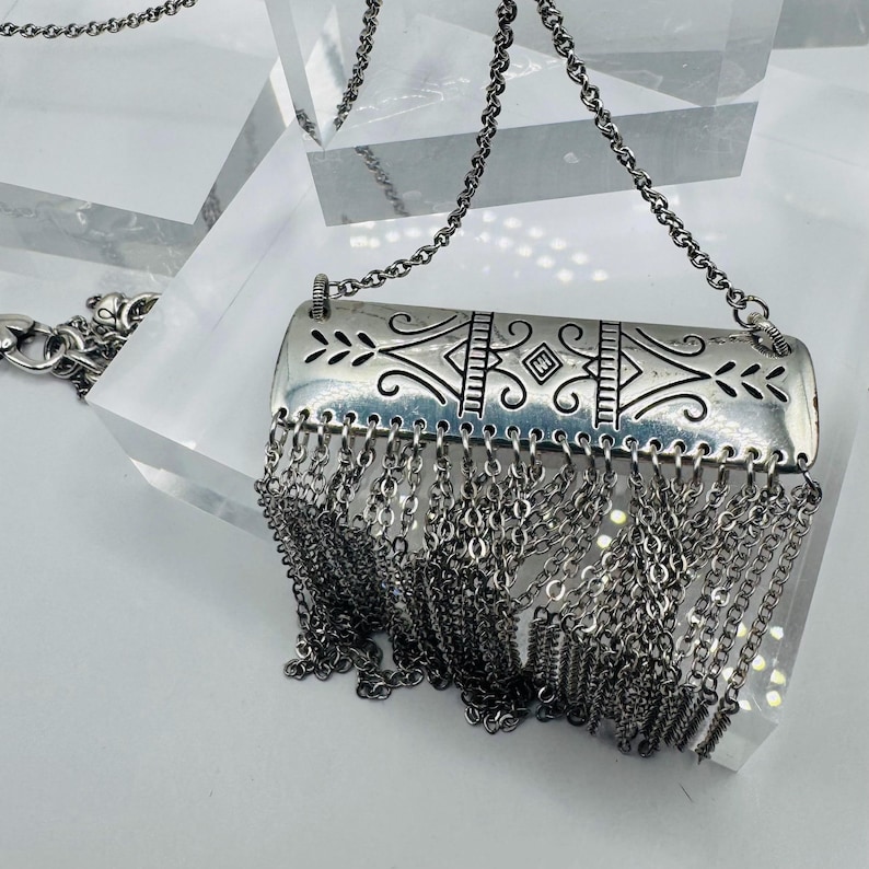 May include: A silver-toned necklace featuring a rectangular metal pendant with engraved symmetrical scrollwork and geometric patterns. A dense fringe of multiple thin silver-toned metal chains hangs from the bottom edge of the pendant.