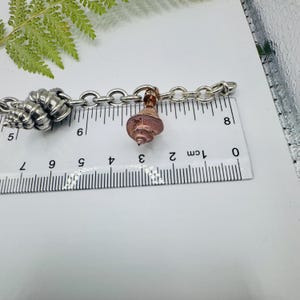 May include: A silver-toned metal chain bracelet featuring a copper-coloured seashell charm and a larger silver seashell charm. A white ruler is positioned beneath the bracelet, showing the copper charm measures approximately 1.3 cm in width.