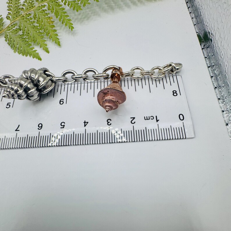 May include: A silver-toned metal chain bracelet featuring a copper-coloured seashell charm and a larger silver seashell charm. A white ruler is positioned beneath the bracelet, showing the copper charm measures approximately 1.3 cm in width.