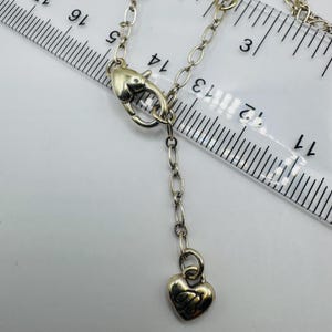 May include: A silver-toned metal necklace chain featuring a heart-shaped lobster clasp and a small heart-shaped charm at the end of an extender chain, positioned next to a clear plastic ruler showing measurements in centimeters.