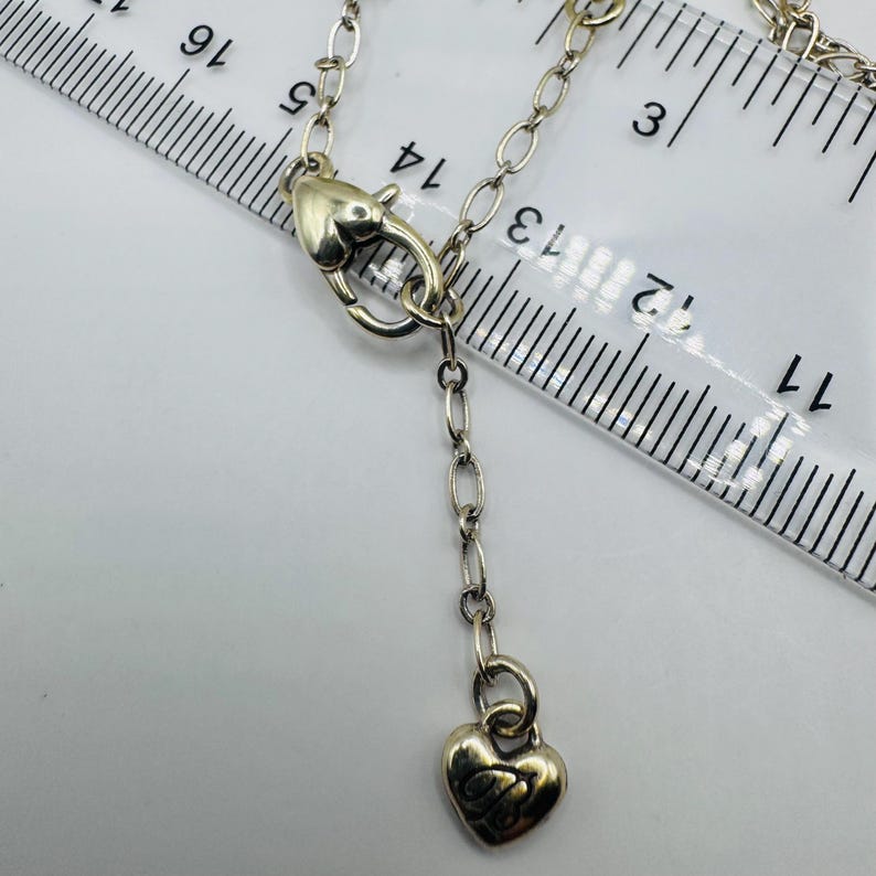 May include: A silver-toned metal necklace chain featuring a heart-shaped lobster clasp and a small heart-shaped charm at the end of an extender chain, positioned next to a clear plastic ruler showing measurements in centimeters.