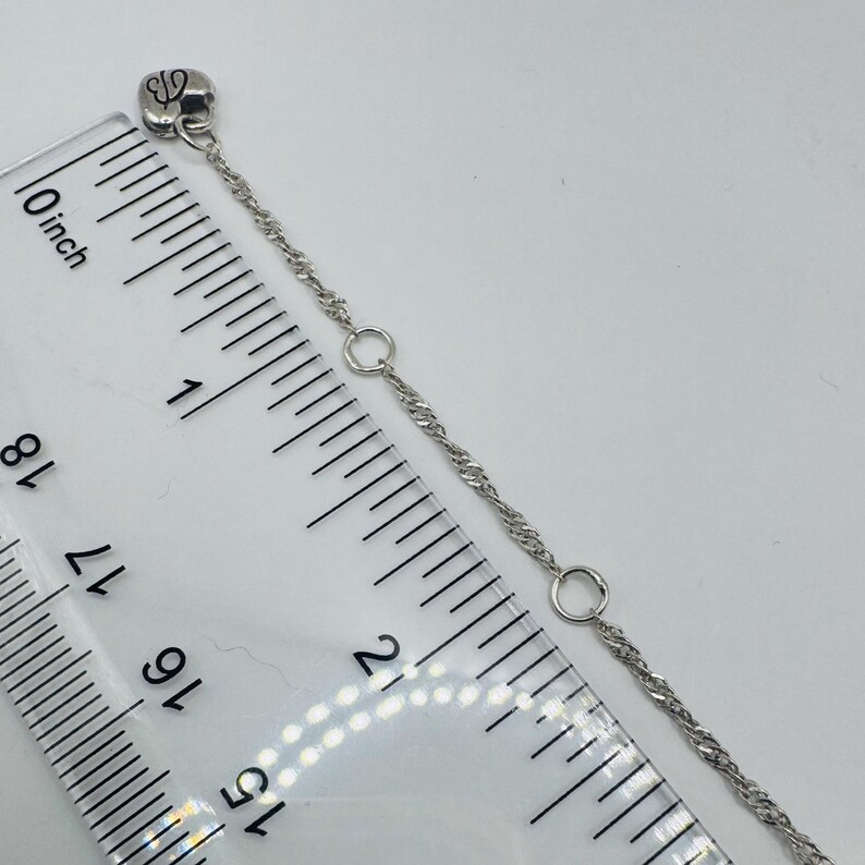 May include: A silver-toned chain with a small heart-shaped charm featuring the letters GB. The chain includes two circular jump rings and is positioned alongside a clear plastic ruler showing a length of approximately 2.5 inches.