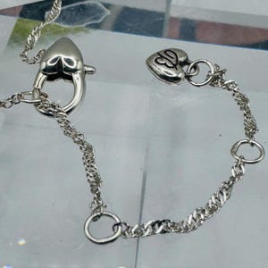 May include: A silver-toned chain necklace featuring a heart-shaped lobster claw clasp and a small heart-shaped charm at the end of the extension chain. The charm is engraved with a stylized letter B. The chain has a twisted, textured link design.