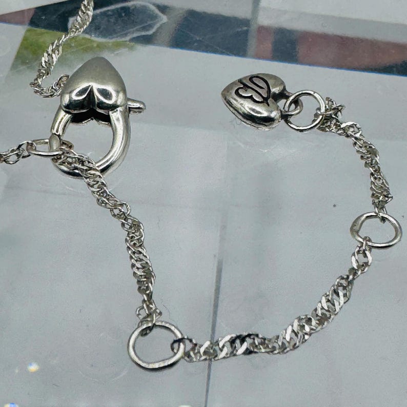May include: A silver-toned chain necklace featuring a heart-shaped lobster claw clasp and a small heart-shaped charm at the end of the extension chain. The charm is engraved with a stylized letter B. The chain has a twisted, textured link design.