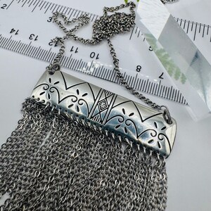 May include: A silver-toned necklace featuring a rectangular metal pendant with engraved scrollwork and geometric patterns, including the letters HH. A dense fringe of fine silver chains hangs from the bottom, measured against a ruler showing 14 inches.