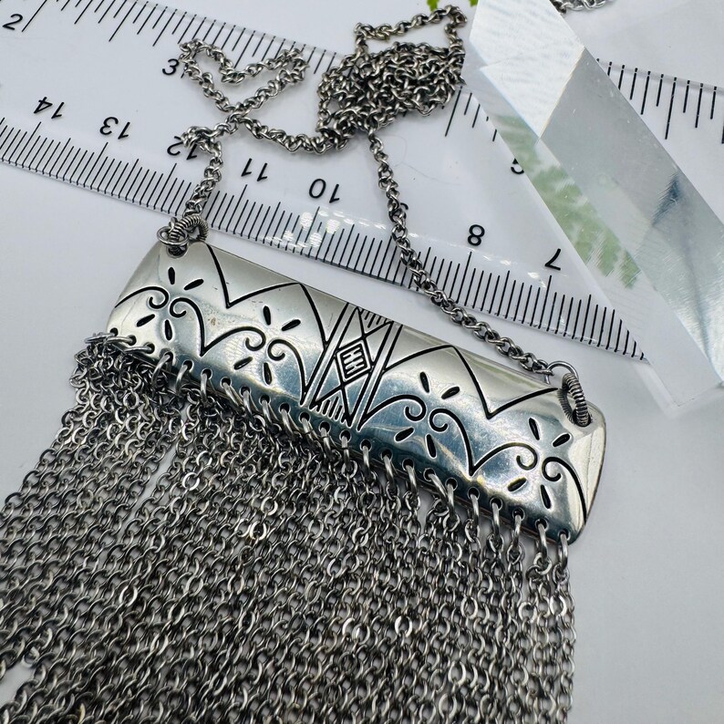May include: A silver-toned necklace featuring a rectangular metal pendant with engraved scrollwork and geometric patterns, including the letters HH. A dense fringe of fine silver chains hangs from the bottom, measured against a ruler showing 14 inches.