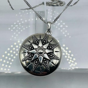 May include: A round silver-toned locket pendant featuring an embossed eight-pointed star design with intricate dotted detailing and a central circular accent, suspended from a delicate silver-toned chain with a polished metallic finish.