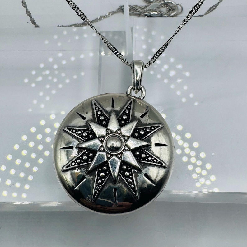May include: A round silver-toned locket pendant featuring an embossed eight-pointed star design with intricate dotted detailing and a central circular accent, suspended from a delicate silver-toned chain with a polished metallic finish.