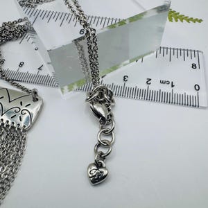 May include: A silver-toned necklace featuring a rectangular pendant with etched patterns and a multi-strand chain fringe. The necklace includes an adjustable chain with a heart-shaped charm, shown next to a ruler measuring approximately 8 inches.