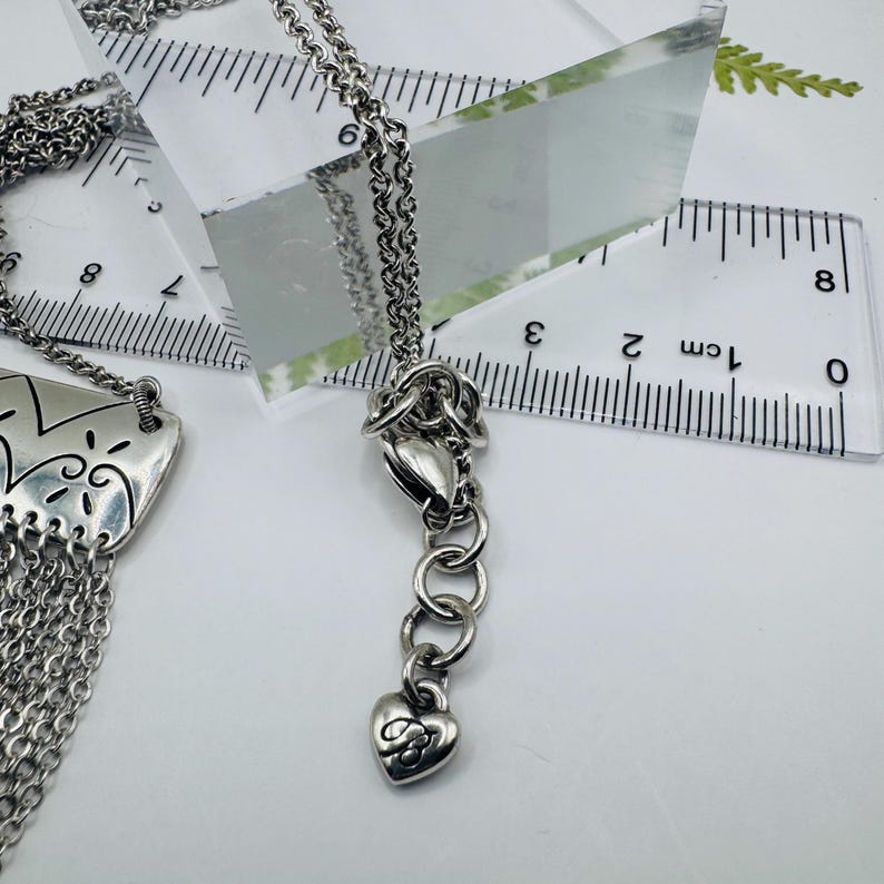 May include: A silver-toned necklace featuring a rectangular pendant with etched patterns and a multi-strand chain fringe. The necklace includes an adjustable chain with a heart-shaped charm, shown next to a ruler measuring approximately 8 inches.