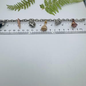 May include: A silver-toned metal charm bracelet featuring various seashell-shaped charms in silver, gold, and copper tones. The bracelet is positioned along a clear plastic ruler, showing a total length of approximately 18 centimetres.