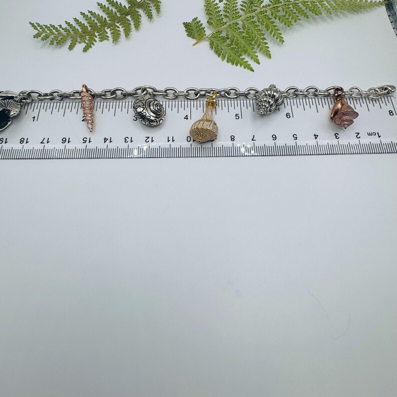 May include: A silver-toned metal charm bracelet featuring various seashell-shaped charms in silver, gold, and copper tones. The bracelet is positioned along a clear plastic ruler, showing a total length of approximately 18 centimetres.