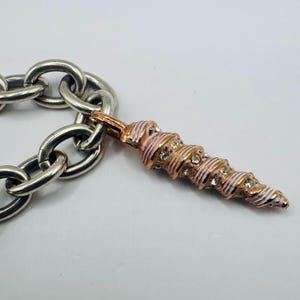 May include: A silver-toned chain link bracelet featuring a single pendant shaped like a spiral seashell. The pendant is finished in rose gold and white enamel, accented with small, clear, round-cut crystals embedded along the spiral ridges.