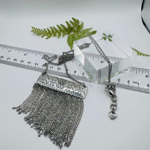 May include: A silver-toned necklace featuring a rectangular metal pendant with engraved floral patterns and a long fringe of delicate chains. A ruler shows the pendant width is approximately 1.5 inches, and a small heart charm is on the clasp.