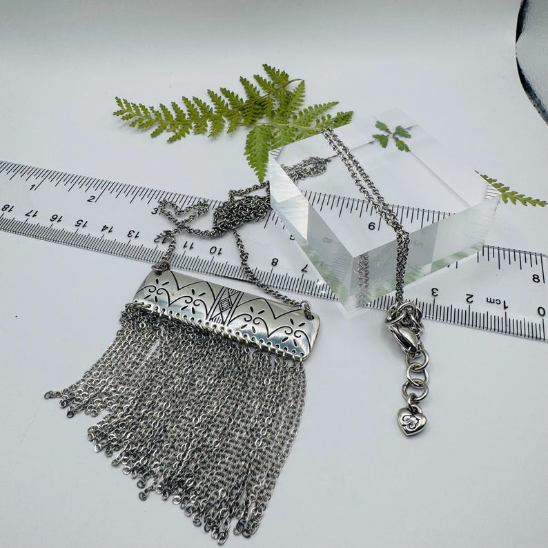 May include: A silver-toned necklace featuring a rectangular metal pendant with engraved floral patterns and a long fringe of delicate chains. A ruler shows the pendant width is approximately 1.5 inches, and a small heart charm is on the clasp.