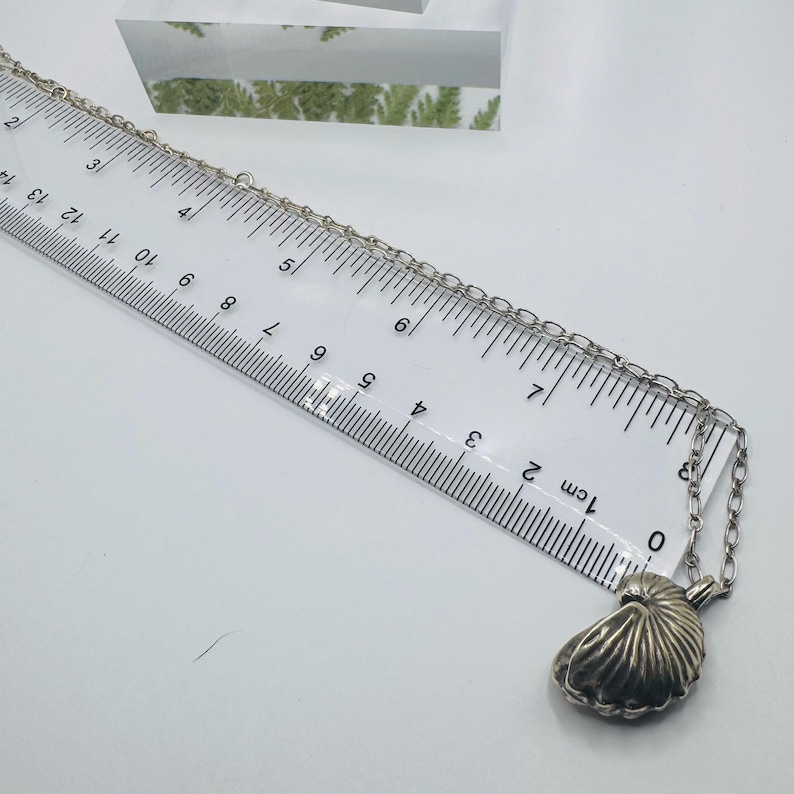 May include: A silver-toned necklace featuring a nautilus shell pendant, positioned alongside a ruler that shows the chain length extending past 14 inches. The pendant has a ribbed, fan-like texture and is attached to a simple metal link chain.