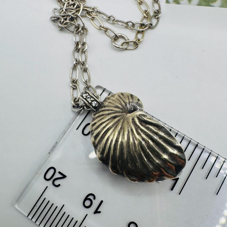 May include: A silver-toned nautilus shell pendant on a matching link chain, positioned next to a clear plastic ruler showing a measurement of approximately 1.25 inches in length. The pendant features a textured, ribbed surface and small clear stones.