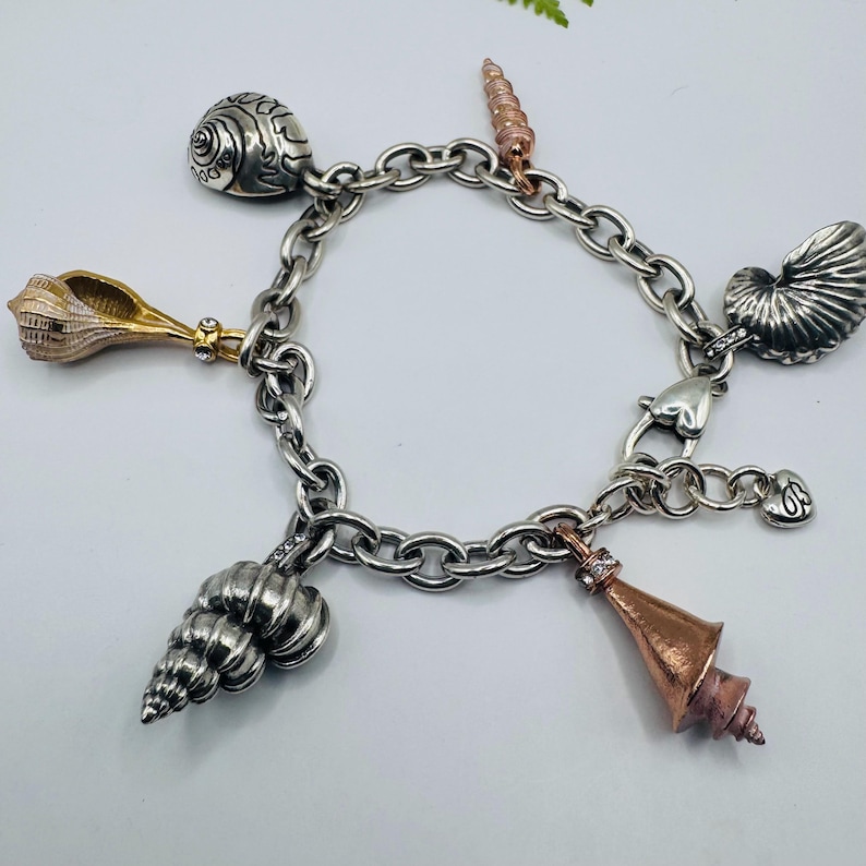 May include: A silver-toned chain link charm bracelet featuring multiple seashell-shaped charms in various metallic finishes, including silver, gold, and copper tones, with small embedded clear stones on several of the shell charms.