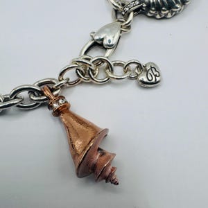 May include: A silver charm bracelet featuring a copper-toned seashell charm with small clear stones at the top. The bracelet includes silver links, heart-shaped charms, and a small heart charm engraved with the letters JB, all in a metallic finish.