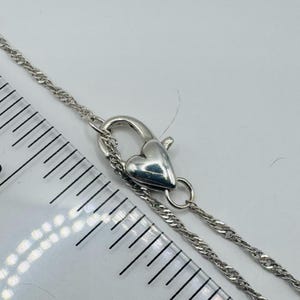 May include: A silver-toned chain necklace featuring a heart-shaped lobster claw clasp. The clasp is positioned next to a clear plastic ruler, showing the heart-shaped mechanism measures approximately 0.5 inches in length and width.