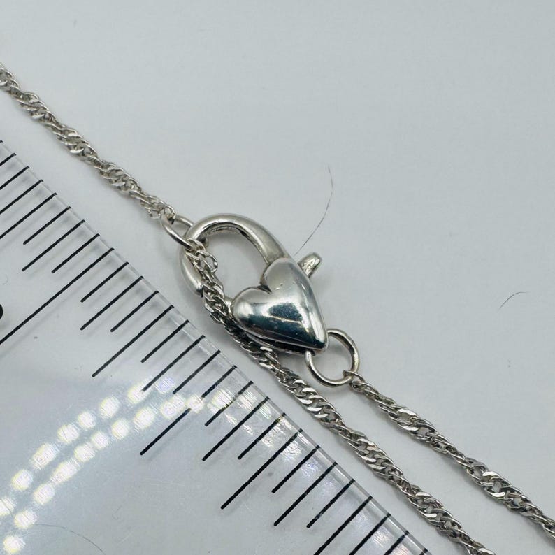 May include: A silver-toned chain necklace featuring a heart-shaped lobster claw clasp. The clasp is positioned next to a clear plastic ruler, showing the heart-shaped mechanism measures approximately 0.5 inches in length and width.