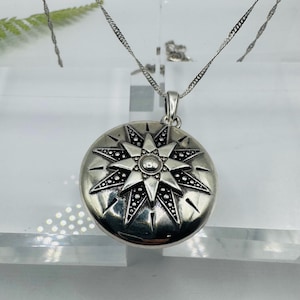 May include: A round silver locket pendant featuring an embossed eight-pointed star design with oxidized black detailing between the rays. The pendant hangs from a thin silver chain, showcasing a polished metallic finish and intricate starburst patterns.