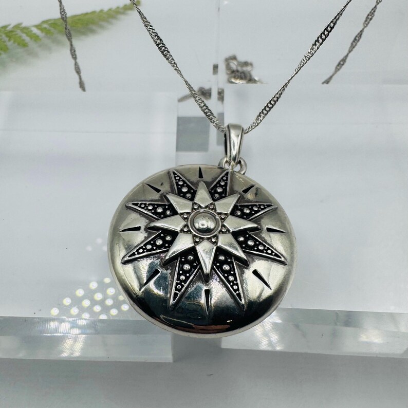 May include: A round silver locket pendant featuring an embossed eight-pointed star design with oxidized black detailing between the rays. The pendant hangs from a thin silver chain, showcasing a polished metallic finish and intricate starburst patterns.