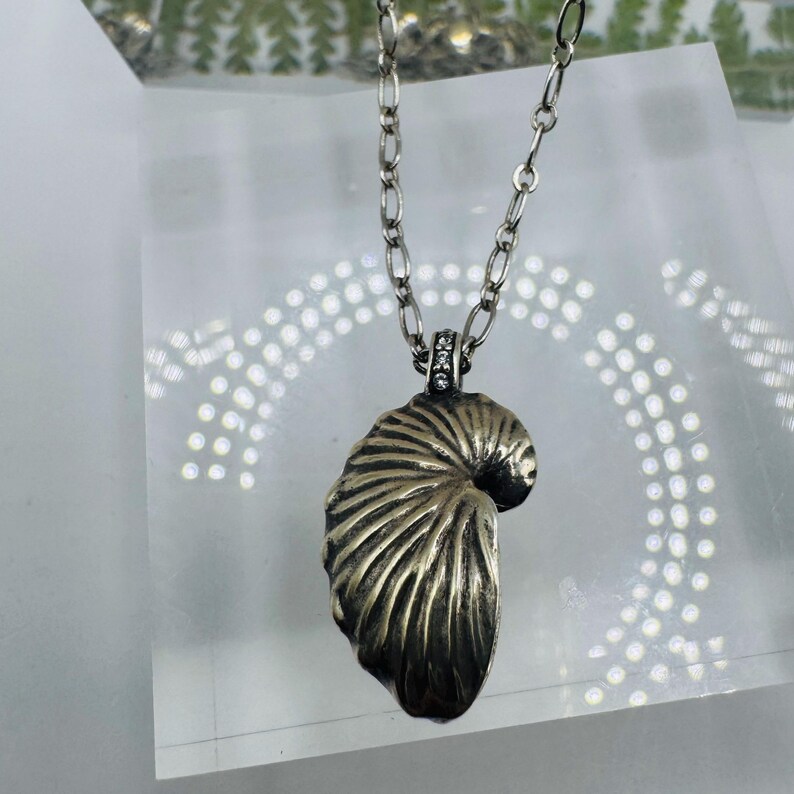 May include: A silver-toned necklace featuring a pendant shaped like a nautilus shell with deep, textured ridges. The pendant is attached to a silver-toned oval link chain and includes a small bail accented with tiny, clear, sparkling stones.