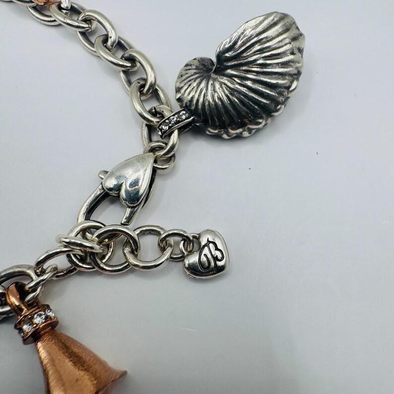 May include: A silver chain link charm bracelet featuring a large textured seashell charm, a heart-shaped clasp, and a small heart-shaped tag with the letters JB engraved. A copper-toned bell-shaped charm with clear crystals is also attached.