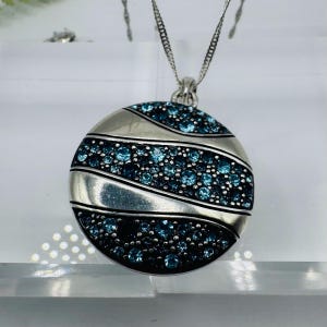 May include: A circular silver pendant suspended from a delicate silver chain. The pendant features three curved, polished silver bands alternating with three sections densely encrusted with small, round, light and dark blue sparkling crystals.