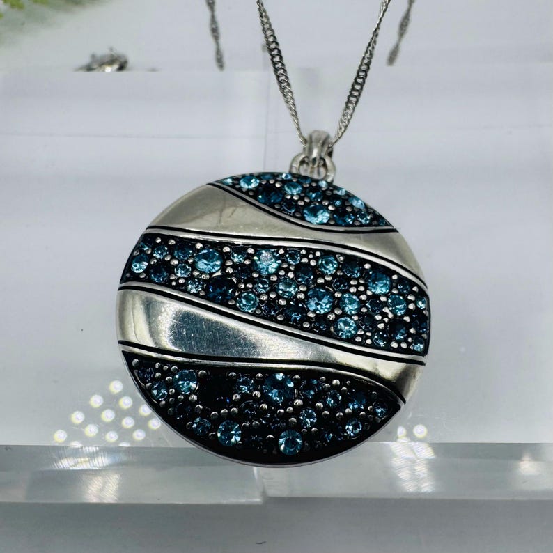 May include: A circular silver pendant suspended from a delicate silver chain. The pendant features three curved, polished silver bands alternating with three sections densely encrusted with small, round, light and dark blue sparkling crystals.